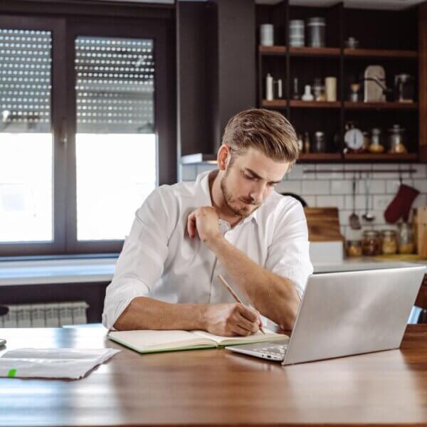 man writes notes in a book while watching laptop screen 