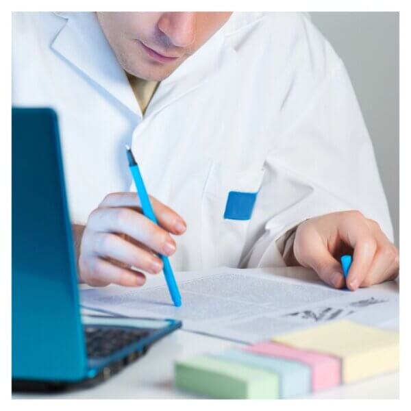Person in a lab coat writing notes next to laptop