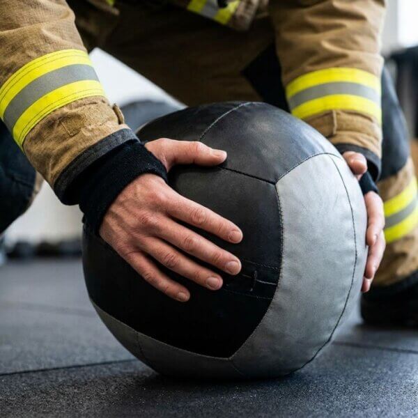 A close-up view shows a firefighter's hands gripping a heavy medicine ball for functional strength training.