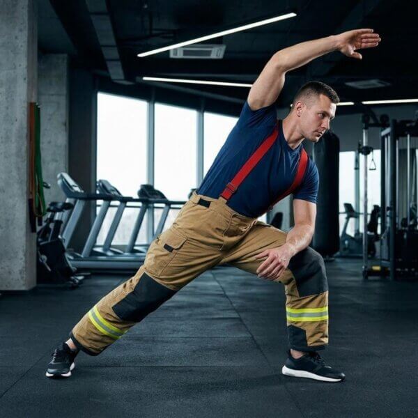 A fit firefighter in athletic wear performs dynamic stretching in a gym for core stability and mobility.